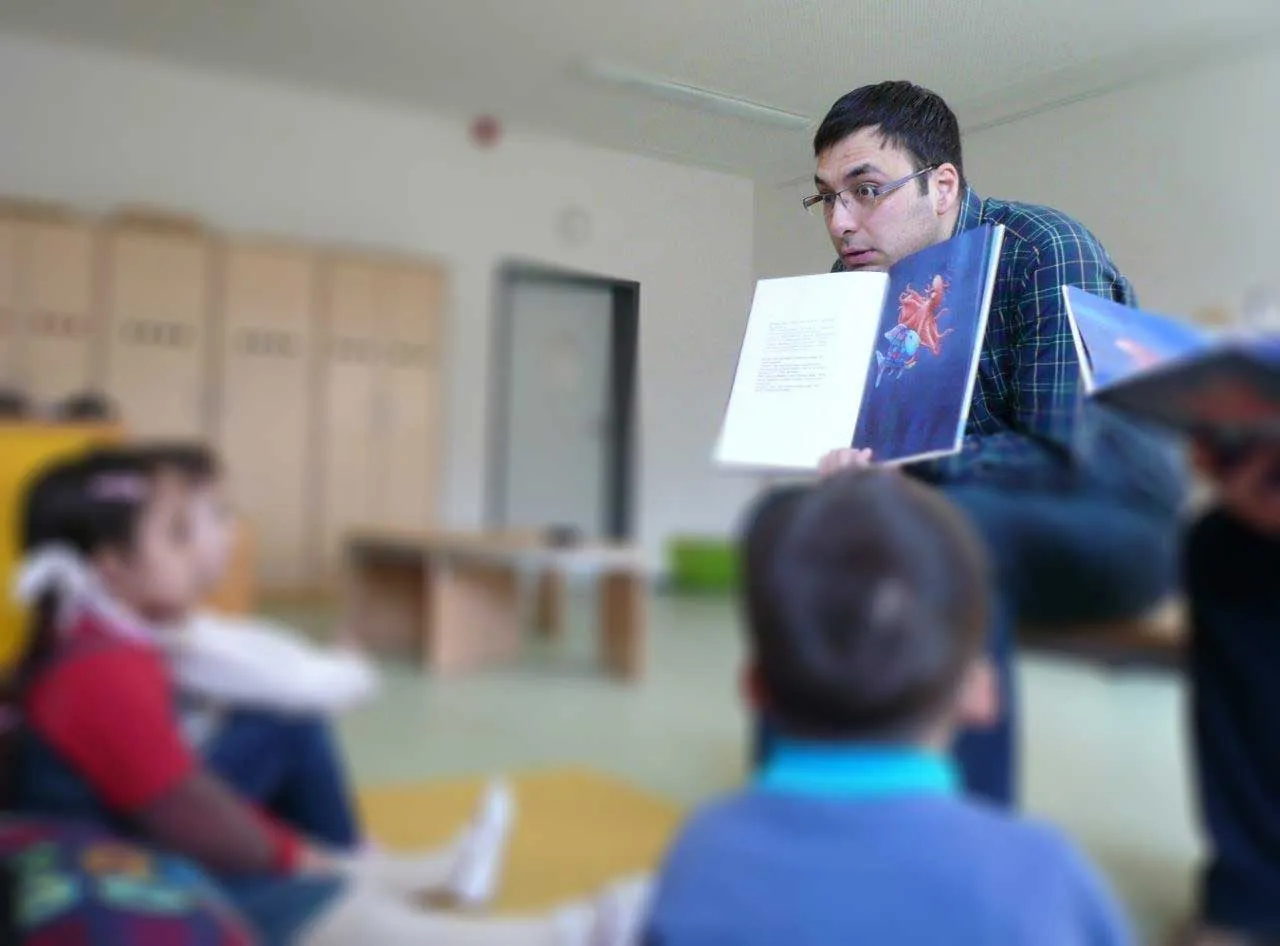 Ömer ließt Kindern in der Grundschule vor und zeigt ihnen in einem Kinderbuch ein Bild.